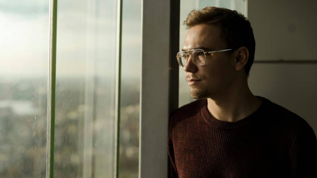 A man in glasses and a red sweater is looking out a large window.
