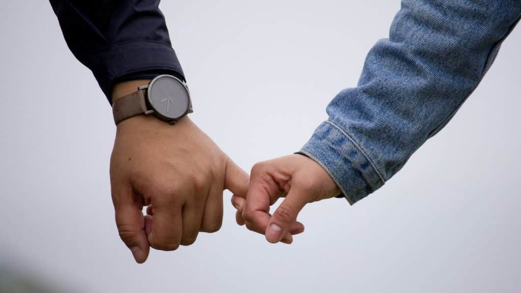 Close-up of a person wearing a watch and a person in a denim jacket holding hands with intertwined pinky fingers.