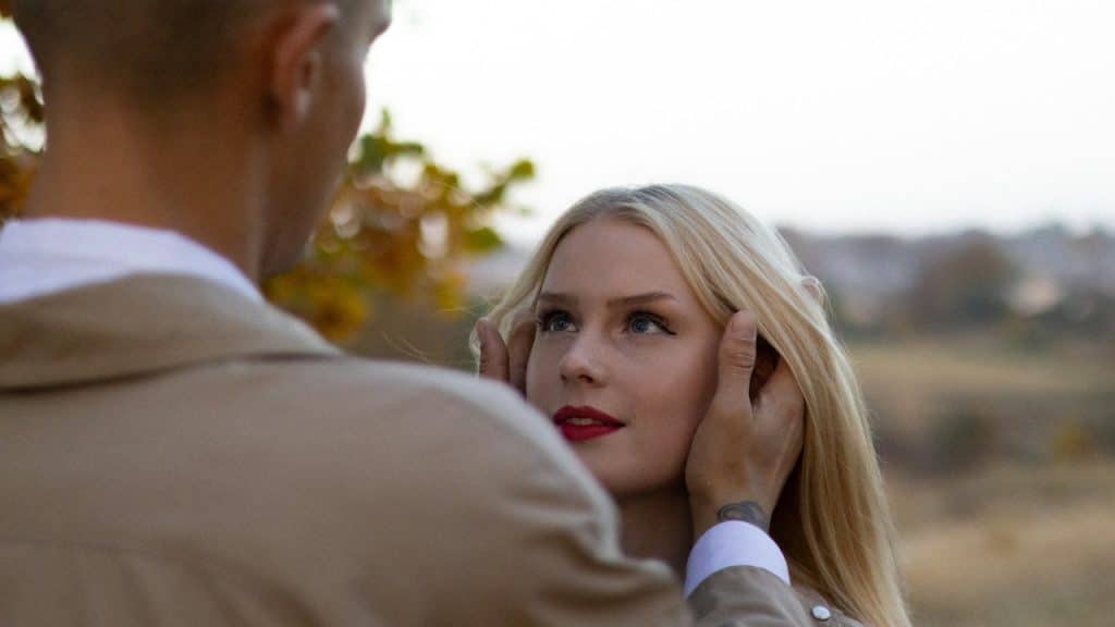 A man gently cups the face of a blonde woman with red lipstick, looking into her eyes outdoors.