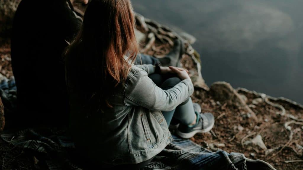 A woman sitting on a blanket near a calm lake, looking out over the water.