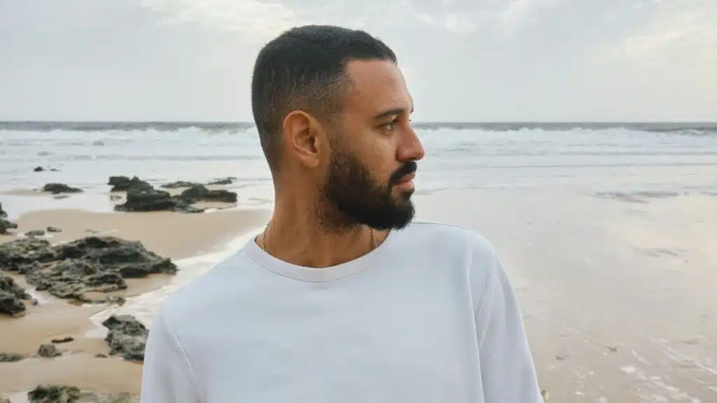 A bearded man in a white shirt stands on a sandy beach looking toward the ocean.