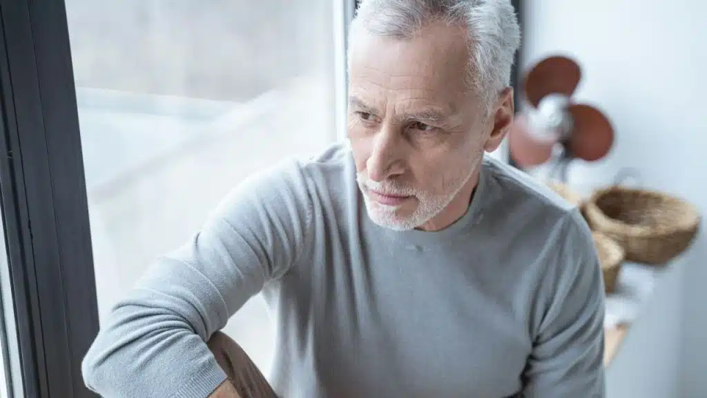 A thoughtful senior man with white hair sits looking out a window.