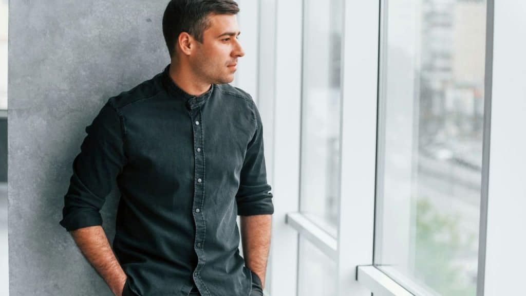 A serious man in a dark shirt stands by a large window, looking out to the side.