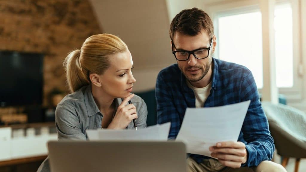 A serious man and woman look at documents while sitting in front of a laptop.
