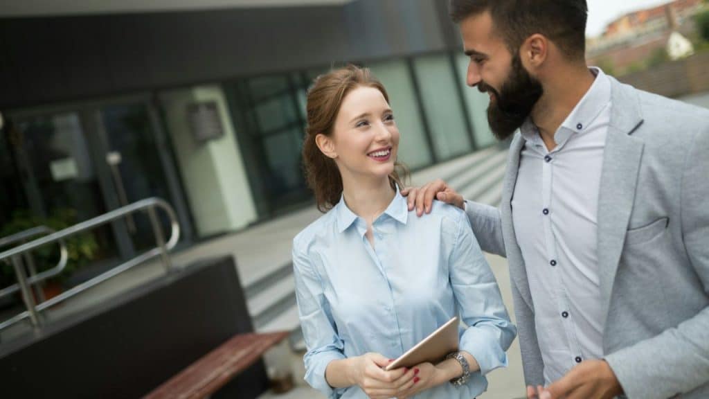 A smiling man with a beard and a woman in a blue shirt walk together outside a building.