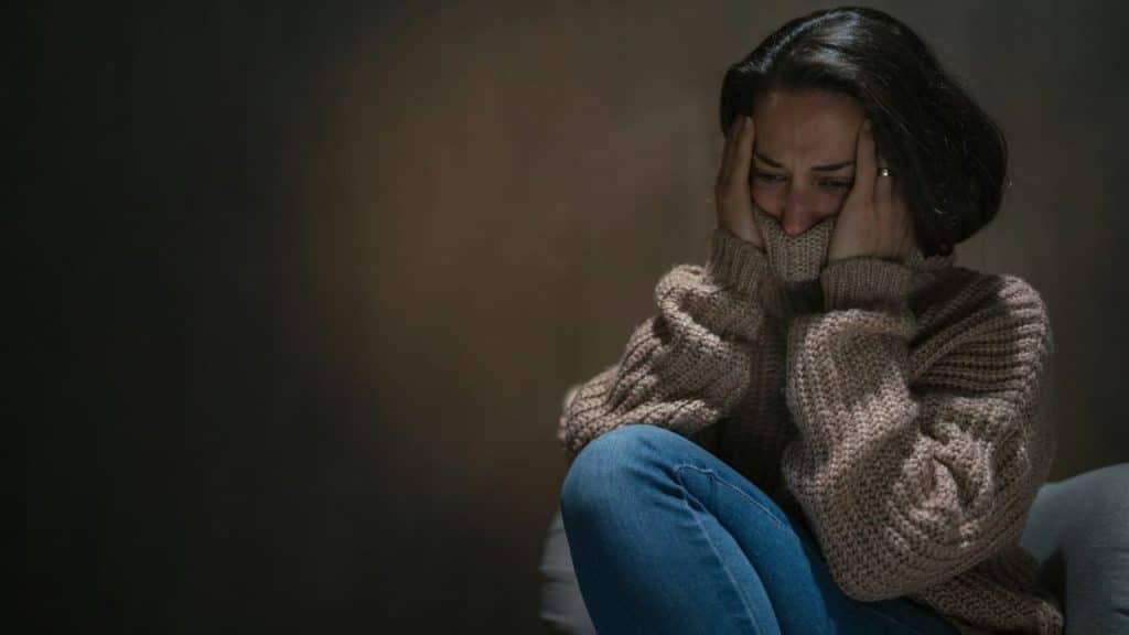 A woman sits huddled, covering her face with her hands and the collar of her sweater.