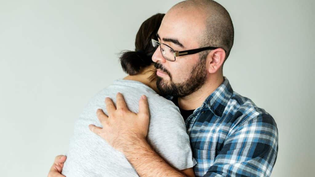 Bald man with a beard and glasses in a plaid shirt hugging another person.