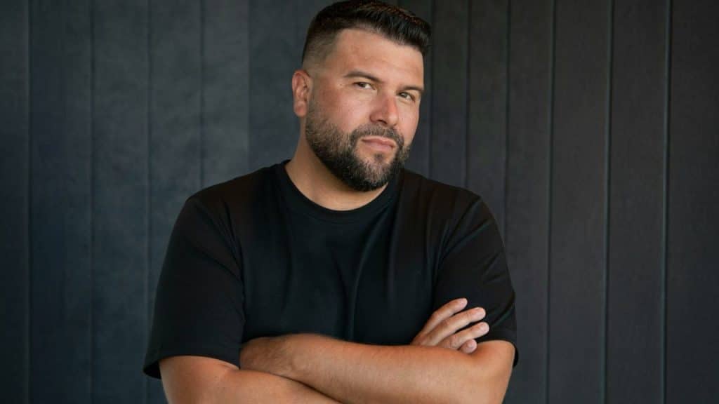 Close-up of a bearded man in a black t-shirt standing with his arms crossed.