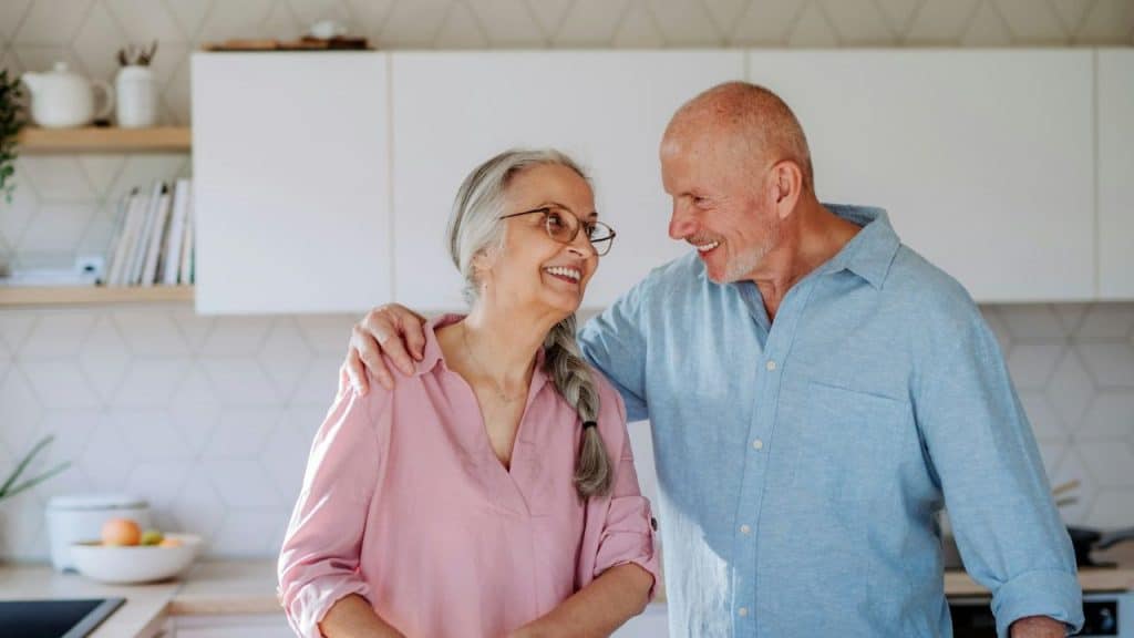 Smiling elderly couple standing close in a modern kitchen, looking at each other affectionately.