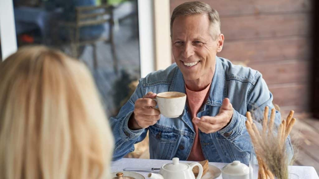 Smiling older man in a denim jacket holding a coffee cup while talking to a blond woman across a table.