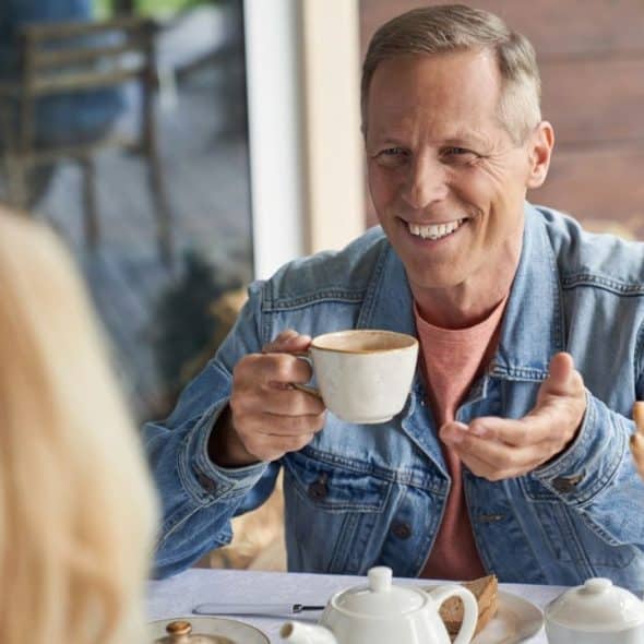 Smiling older man in a denim jacket holding a coffee cup while talking to a blond woman across a table.