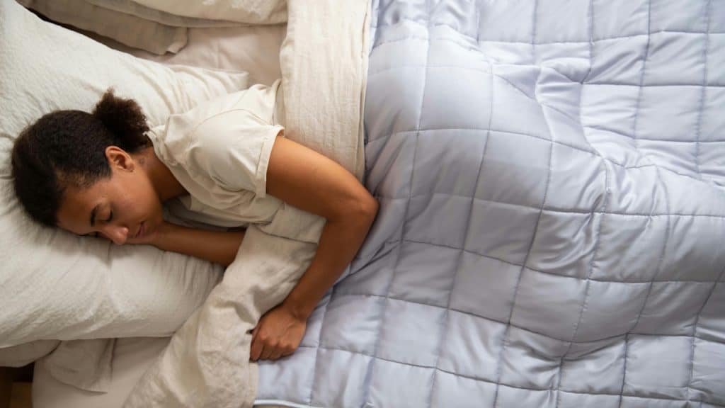 An overhead view of a woman sleeping peacefully under a light blue, quilted weighted blanket in a bed with soft pillows.