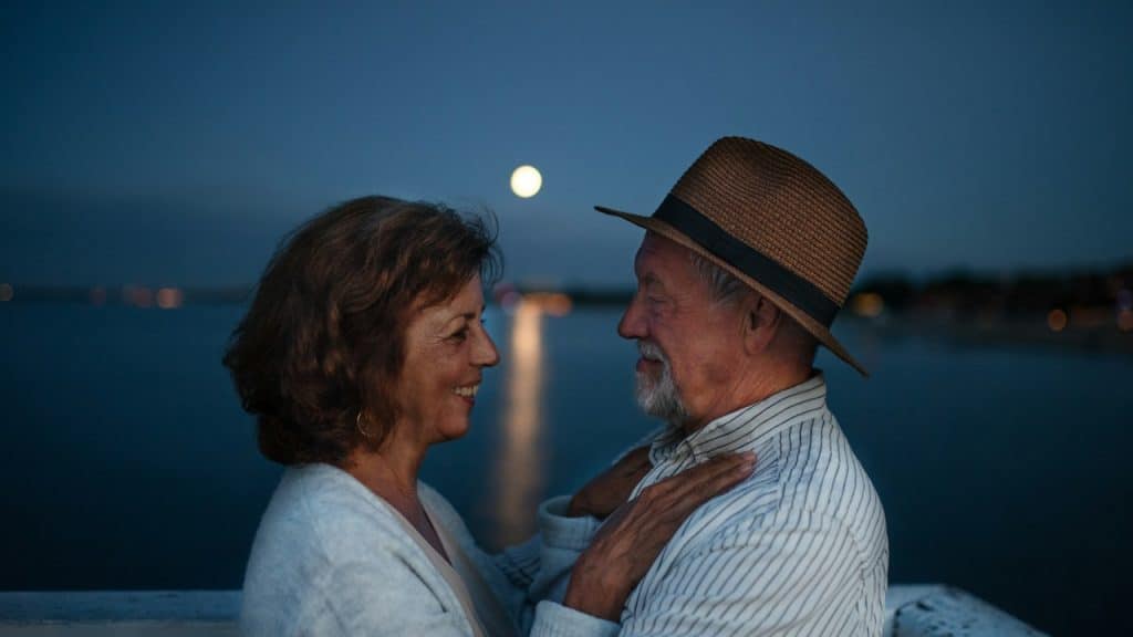 A happy couple hugging outdoors on a pier by the sea at dusk looking at each other.