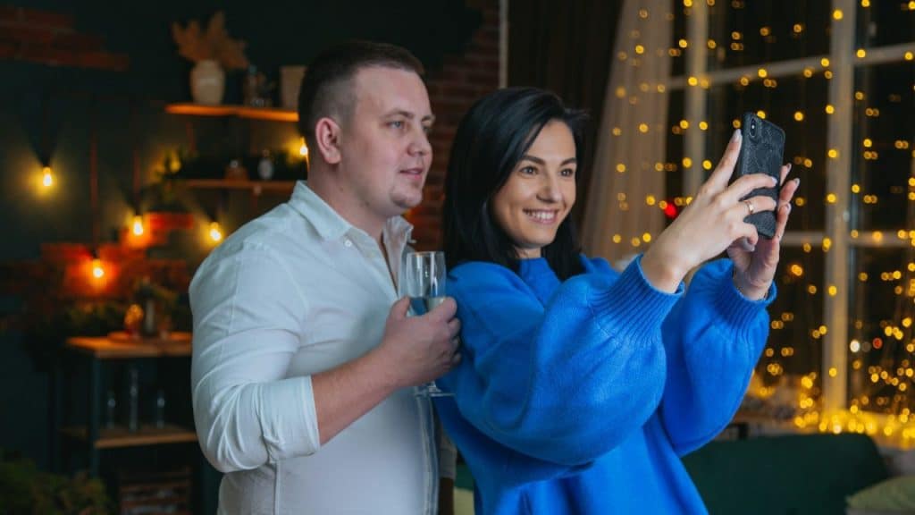 A couple stands in a dimly lit room with string lights, taking a photo with a phone.