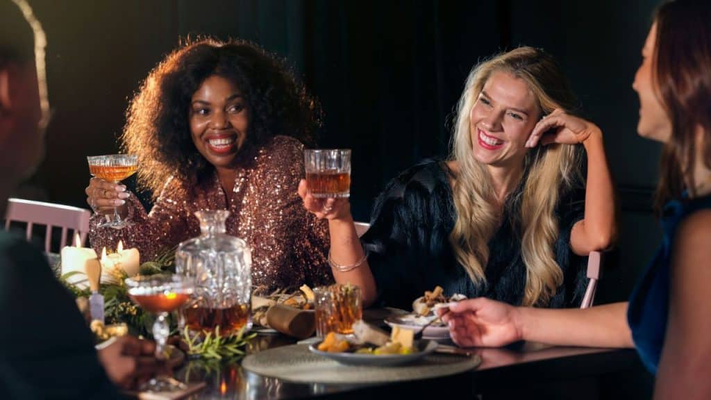 Two women smile while holding drinks at a dimly lit dinner table with friends.