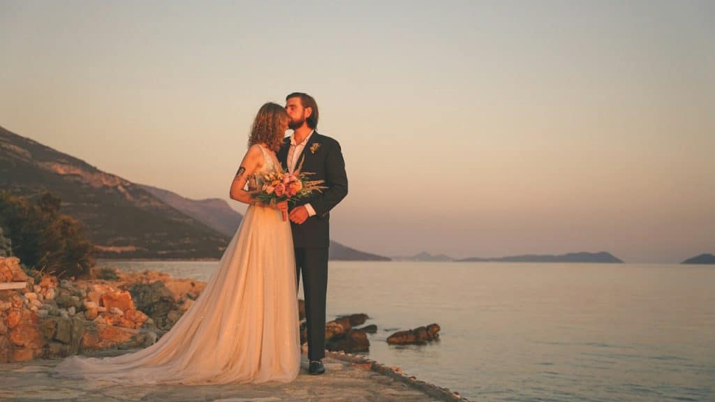 A newly married couple by the seaside where the groom is kissing the brideβs forehead.