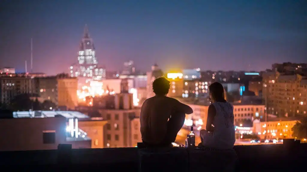 A man and a woman chilling on a rooftop in front of high-rise buildings.