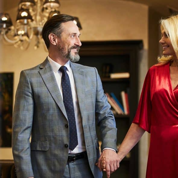 A well-dressed, smiling couple holds hands in an elegantly furnished room.