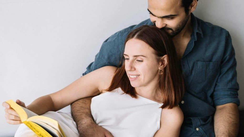 A couple sitting together while the woman reads a book and the man embraces her.