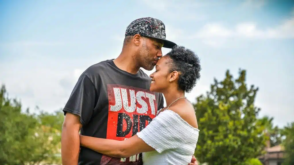 A man in a baseball cap leans in to kiss a smiling woman on the forehead as they embrace outdoors.