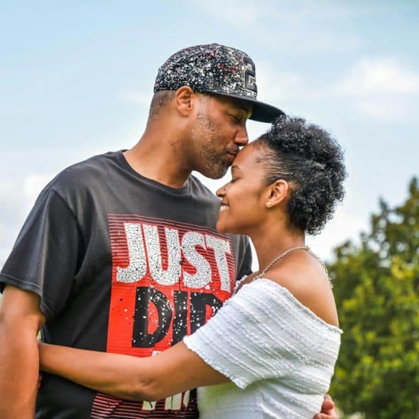 A man in a baseball cap leans in to kiss a smiling woman on the forehead as they embrace outdoors.