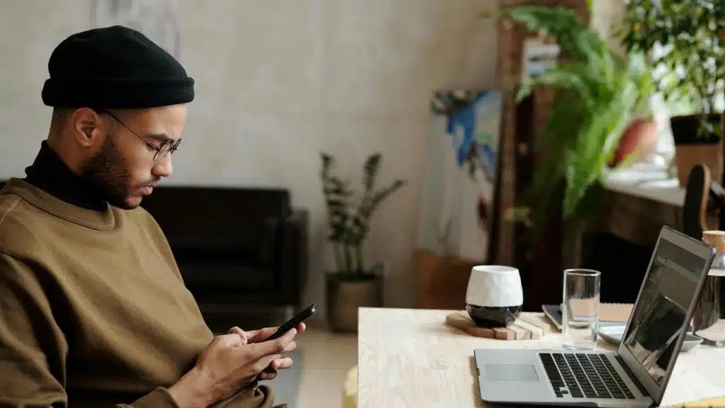 A man sitting at a desk using his phone beside a laptop.