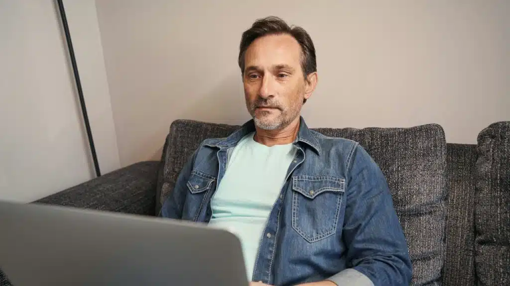 A Man in a denim jacket sitting on a sofa using a laptop.