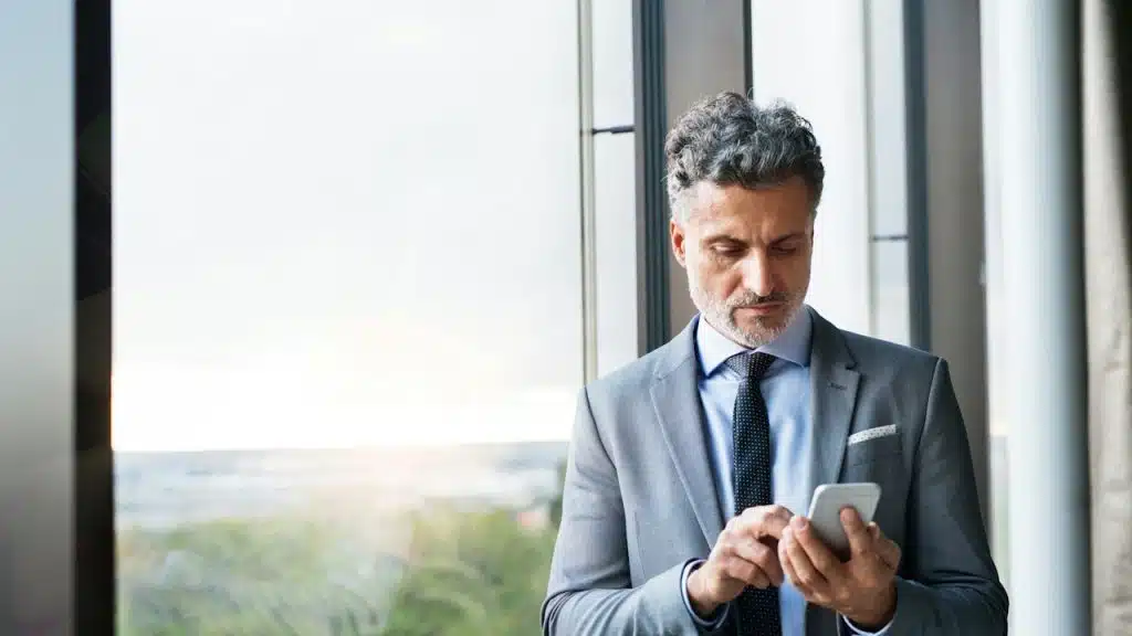 A mature businessman in a light grey suit and tie standing by a large window, intently using a smartphone.