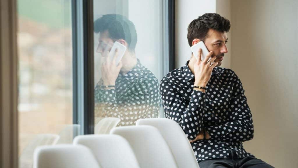 A man in a black and white polka dot shirt sits by a window, talking on a white smartphone.