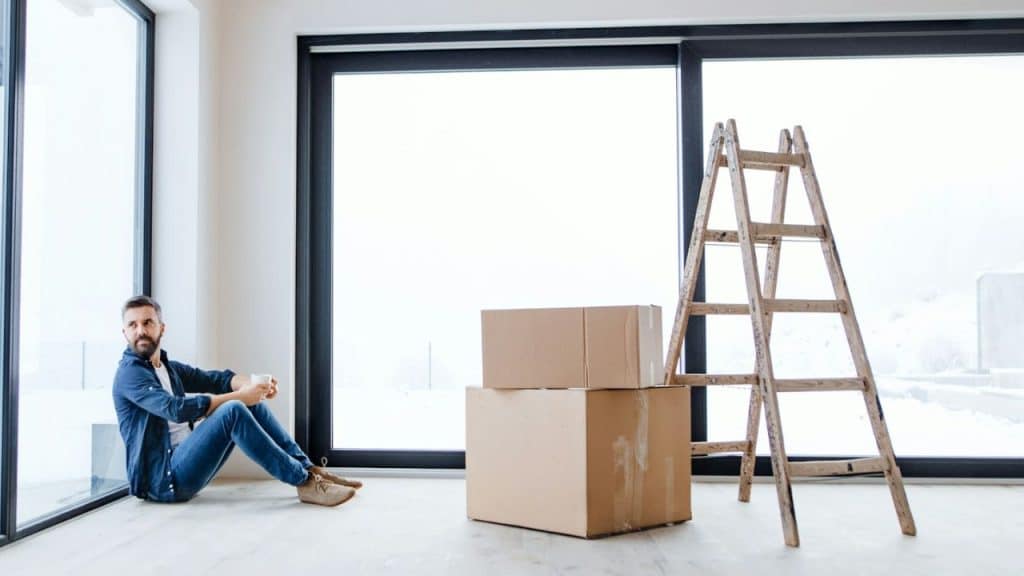 A man sits alone on the floor of an empty room near a large window, with cardboard boxes and a ladder nearby.