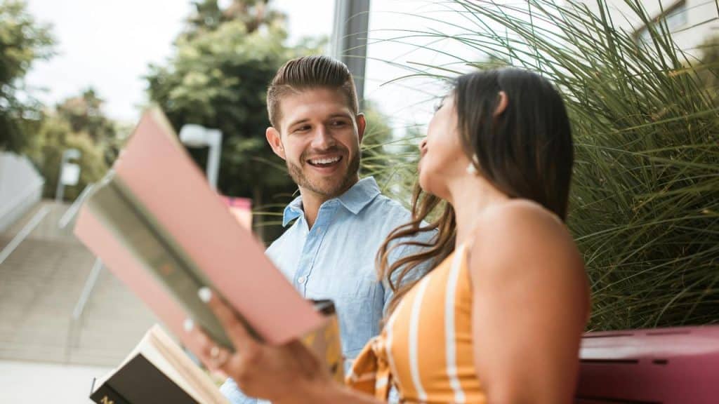 A man and woman sitting outside laughing while holding books.