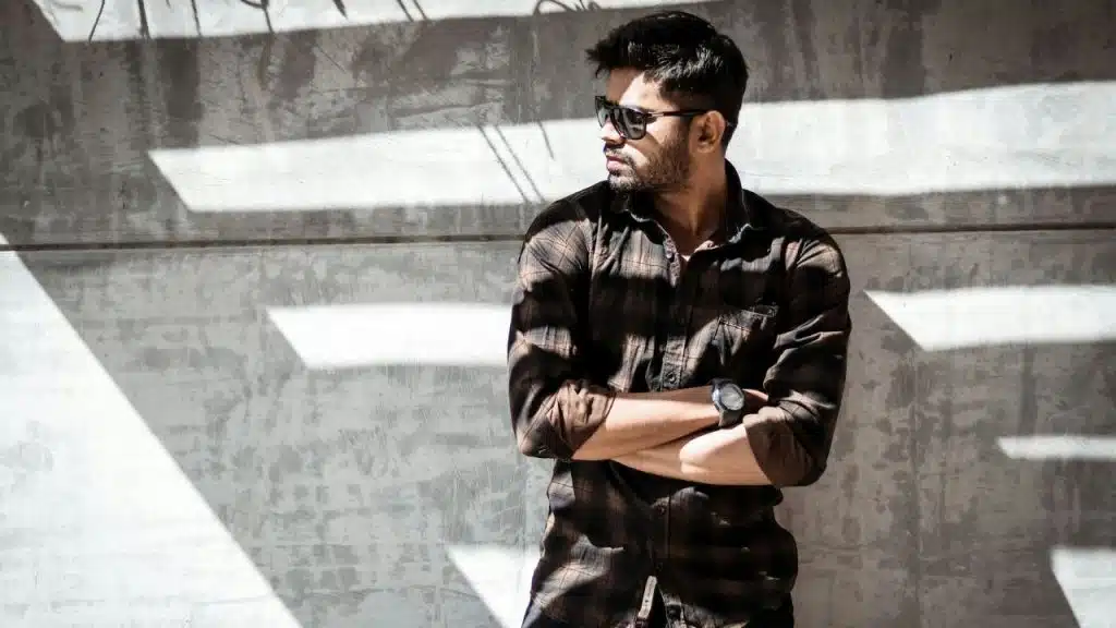 A man in a plaid shirt and sunglasses stands in the sun against a concrete wall with his arms crossed.