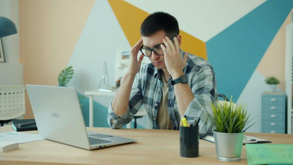 Man in plaid shirt holding his temples, sitting at a desk with a laptop.