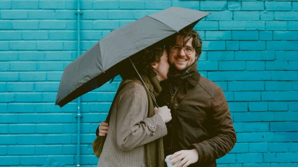 Couple smiling and embracing under an umbrella in front of a blue brick wall.