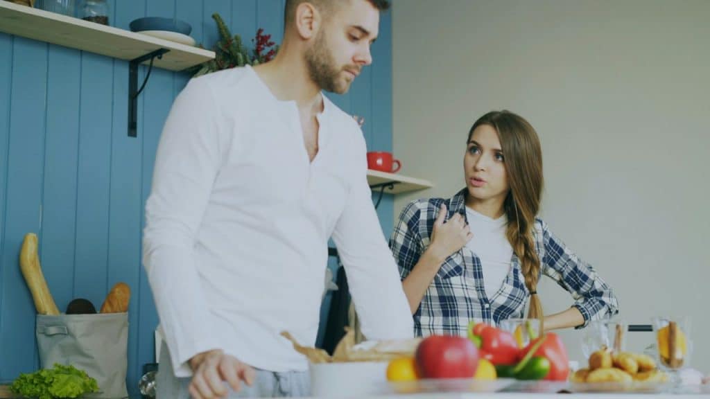 A man and woman are standing in a kitchen, talking around a counter with food on it.