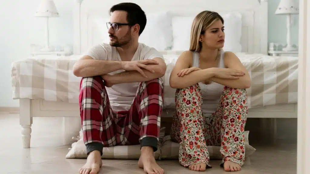 A man in plaid pajama pants and a woman in floral pajama pants sit back-to-back on the floor next to a bed.