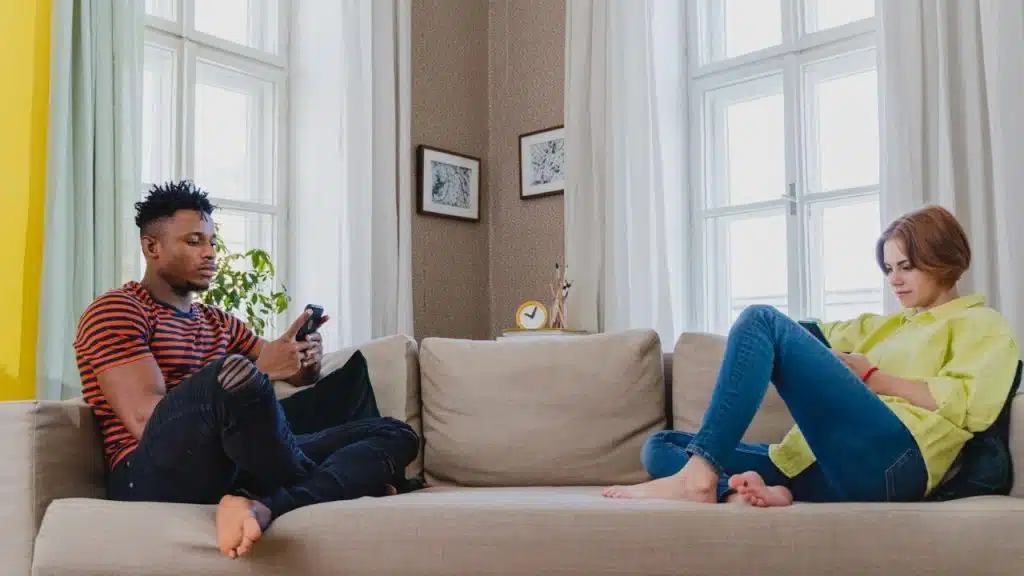 A man and woman sit far apart on a couch, each looking down at their cell phone.
