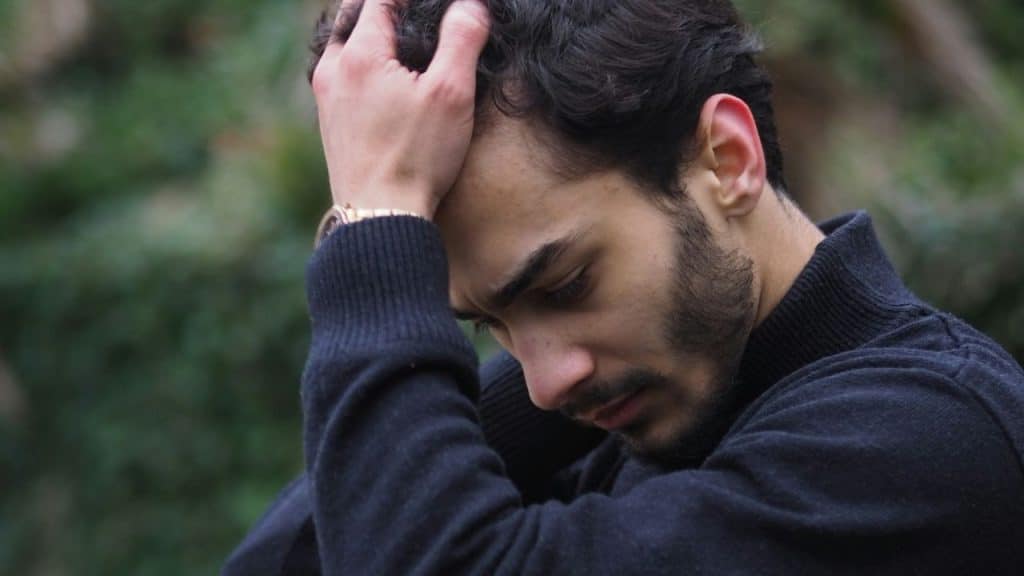 A bearded man with a gold watch and black sweater holds his head in his hands outdoors.