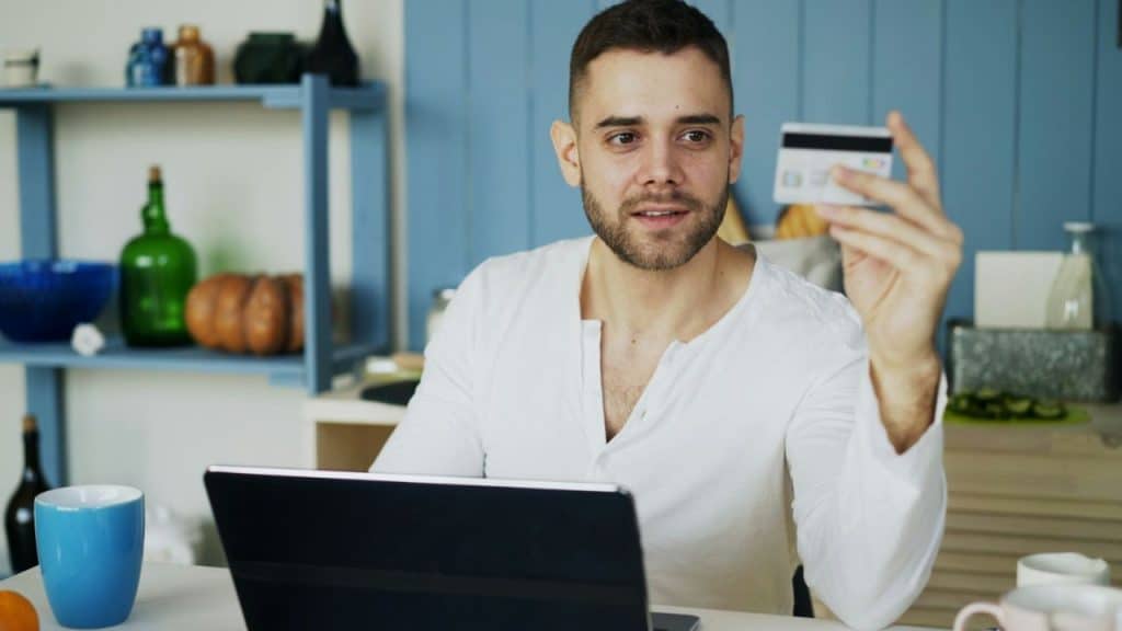Man with a beard sits at a laptop, holding up a credit card.