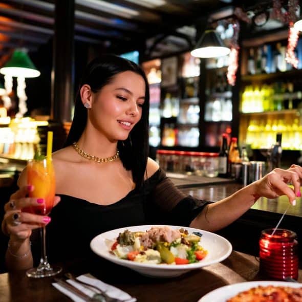 A woman dining at a restaurant while holding a cocktail and smiling at her meal.