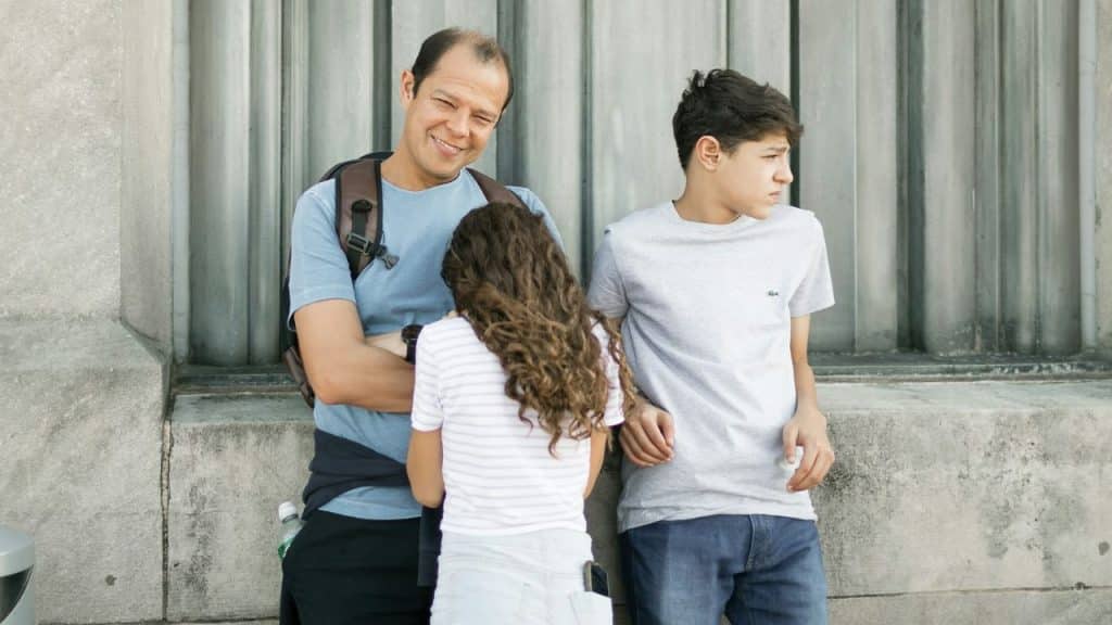 A smiling man with a backpack stands with two children against a gray stone wall, one girl facing away and one boy looking to the side.