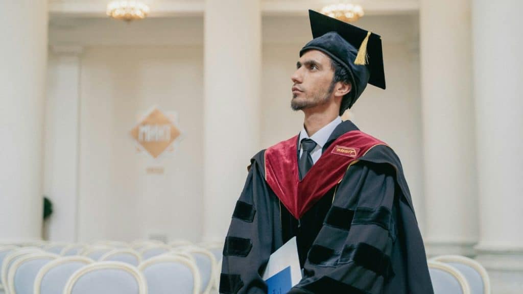 A graduate in a cap and gown stands in a large hall with rows of empty chairs.