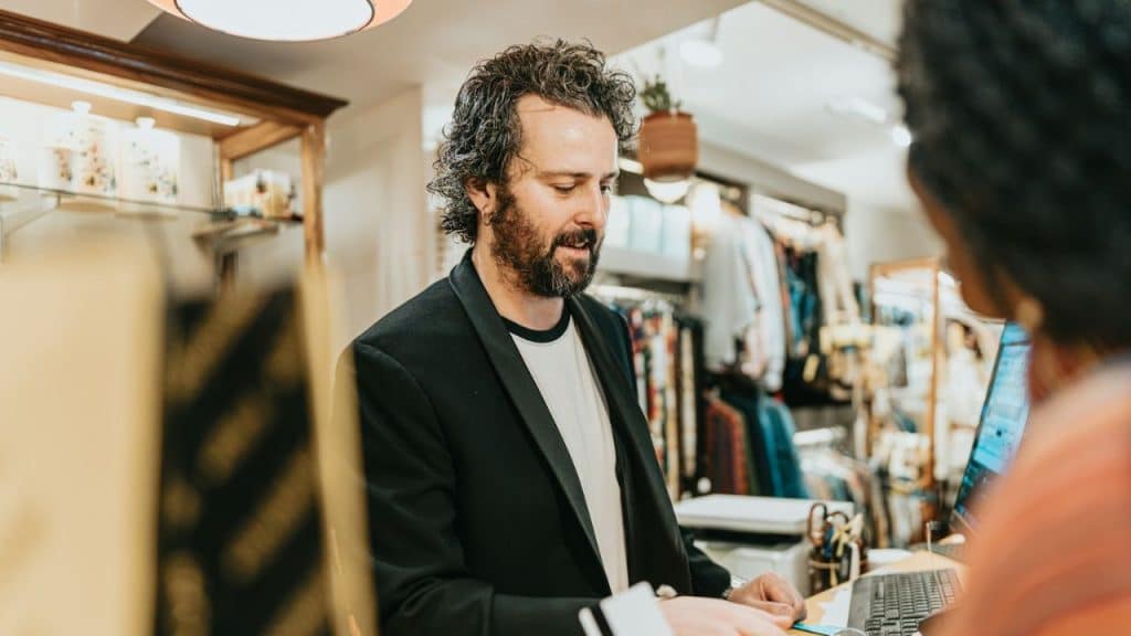 Man with beard, black blazer, and white shirt helping a customer at a store counter.