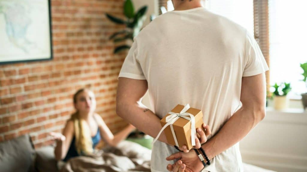 Man hiding a small gift box behind his back, facing a blurry woman on a couch.