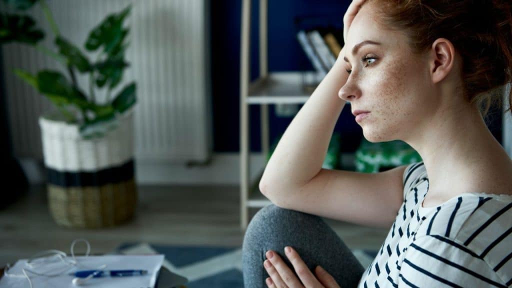 Woman with freckles sitting indoors, resting her head on her hand with a pensive look.