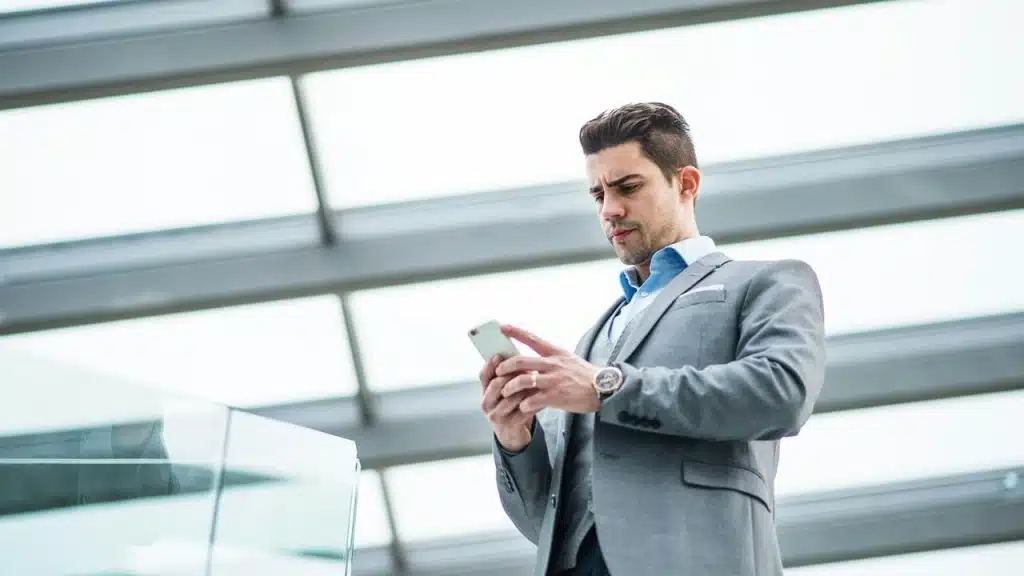 A man in a gray suit looking at his smartphone in a modern glass building.