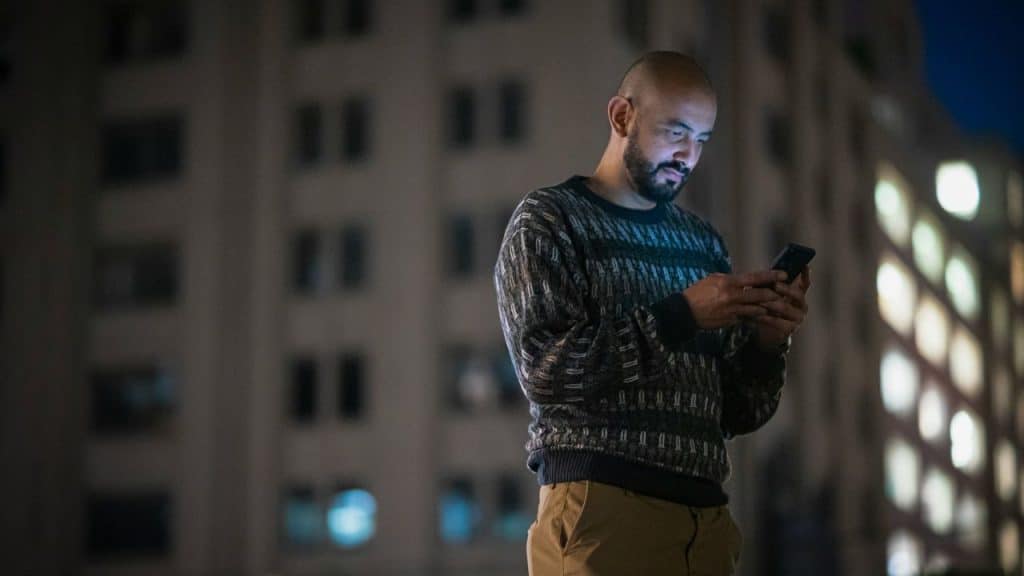 A balding man with a beard is standing outdoors at night, looking intently at his illuminated smartphone.