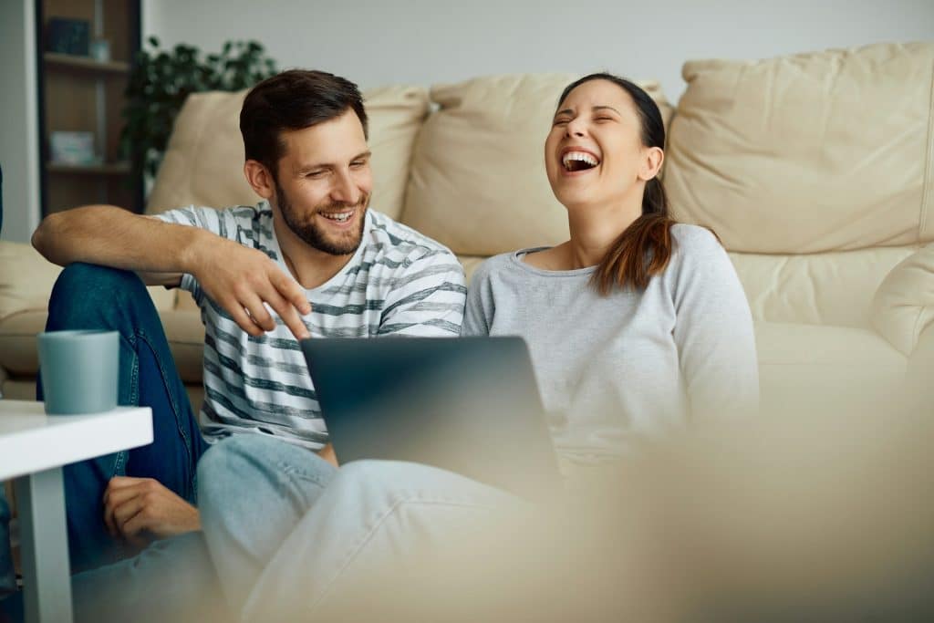 The couple is laughing while looking at the laptop and hanging out in the living room.