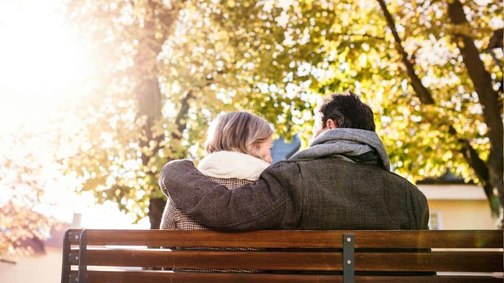 A mature couple sitting on a wooden park bench with the man's arm around the woman, facing a sunlit autumn tree.