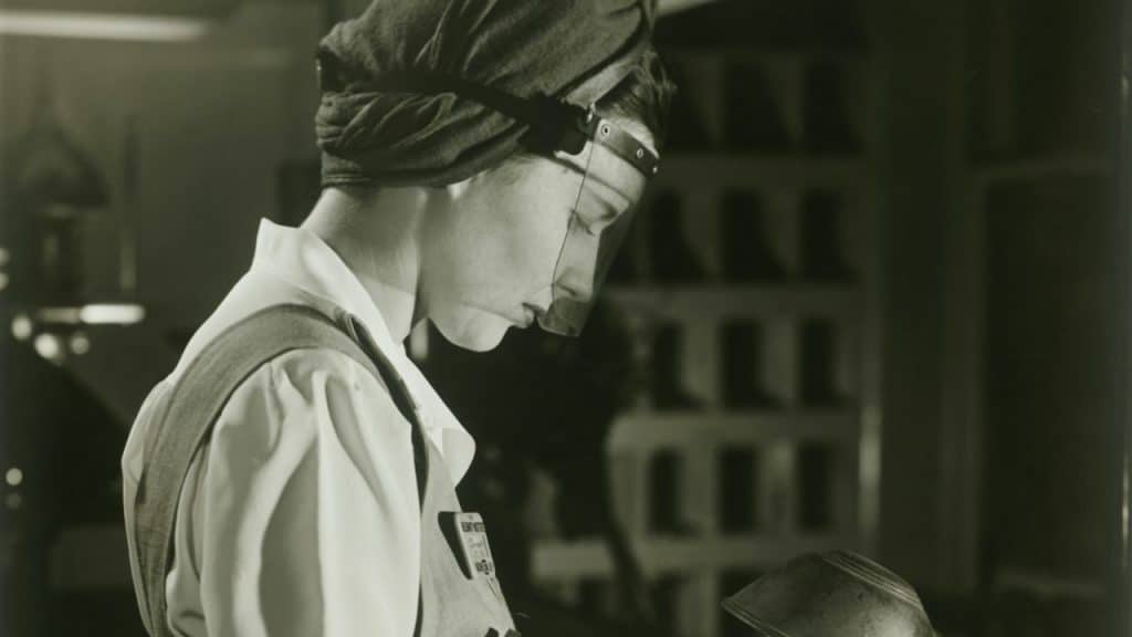 A black and white profile photo of a woman in factory work clothes, wearing a hair wrap and face shield.
